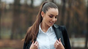 A photo of a beautiful woman with a ponytail. She is wearing a white shirt and a black blazer. She has her eyes closed and is smiling. The background is blurred and consists of trees.