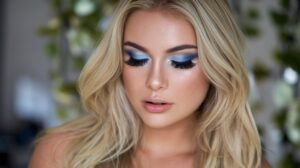 A photo of a stunning blonde woman with blue eyes. She is wearing makeup with blue eyeshadow and false lashes. Her eyes are the focus of the photo. The background is blurred and contains greenery.