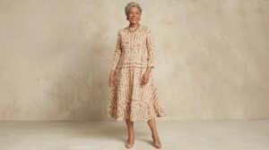 A photo of a woman over 50 wearing a beige midi dress with a unique pattern of abstract shapes. The dress has a modest neckline and a flattering fit. She accessorizes with a gold necklace and a pair of beige pumps. The background is a beige wall.