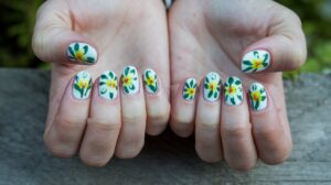 A photo of a woman's hands with white spring nails. The nails have green and yellow flowers with green leaves. The nails have a glossy finish. The hands are placed on a wooden surface. The background is blurred and contains greenery.