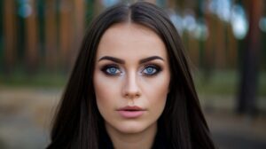 A photo of a woman with blue eyes. She has long, dark hair and is wearing makeup. Her eyes are the main focus of the photo, with a strong emphasis on the blue hue. The background is blurred and consists of a forest with tall trees.