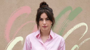 A photo of a woman with dark hair wearing a pink shirt. She has a top knot hairstyle. The background is a beige wall. There are flourishes of pink, green, and white paint on the wall.