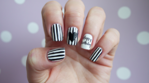 A photo of a person's hand with black and white striped nails. There is a black heart and the text "Happy birthday" painted on the nails. The background is a light purple wall with white polka dots.