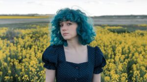 A photo of a woman with curly hair. She has dark blue hair and is wearing a dark blue dress with white polka dots. The woman is standing in a field of yellow flowers. The background is vast and open, with more yellow flowers and a clear sky.