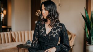 A woman with dark hair is wearing a hairstyle for a date night. She has styled her hair with loose waves, falling gently over her shoulders. She wears a black dress with a v-neck and a floral pattern. The dress is paired with black heels and a black clutch. She is standing in a room with a beige couch, a table, and a plant. The room has a warm lighting.