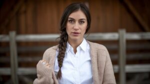 A photo of a woman with long, dark hair wearing a fishtail braid. She is wearing a white shirt and a beige cardigan. The background is a wooden structure. The lighting is soft.