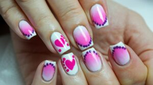 A photo of a trendy nail art design. The nails are painted with a gradient of pink to purple, with white tips. There are small pink and purple polka dots and stripes on the nails. There is a pink and purple heart on the middle finger. The nails are manicured and have a glossy finish.