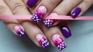 A photo of trendy nail art. The nails have a gradient of pink to purple with white polka dots and a few small pink hearts. There is a pink ribbon wrapped around the middle of the nails. The nails have a glossy finish. The background is a light blue mat.