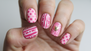 A photo of a hand with a pink nail design. The nails are adorned with various pink patterns, including polka dots, stripes, and swirls. There is also a small gold heart on one of the nails. The background is white.