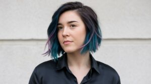 A medium shot of a woman with short hair. She has dark blue hair with purple and pink streaks. She is wearing a black shirt. The background is a white wall.