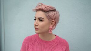 A photo of a woman with short hair wearing a pink sweater. Her hair is styled in a cute, playful manner, with a few strands sticking up in the front. She has pink hair and wears pink earrings that match her sweater. The background is a light blue wall.