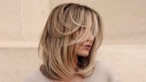 A photo of a woman with a layered hairstyle. Her hair is medium length and has multiple layers, with the top layer being the shortest. The hair is sleek and straight, with a side part. The woman is wearing a beige sweater and a necklace. The background is a beige wall.