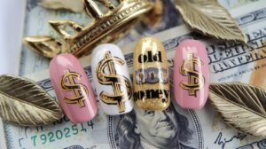 A photo of old money-inspired nails with a vintage touch. The nails have a gold theme, with a dollar sign and the text "Old Money". There are also leaves and a crown. The nails are placed on a white background.